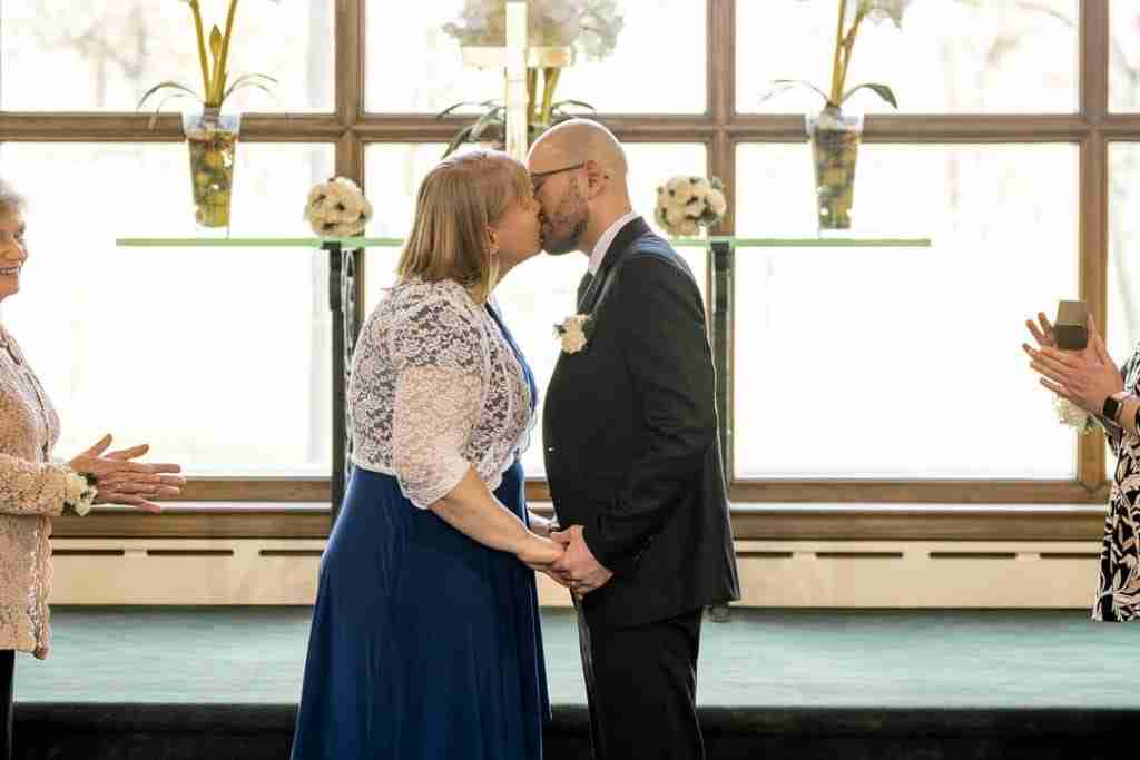 A couple kissing during a wedding ceremony while holding hands, with onlookers clapping in the background.