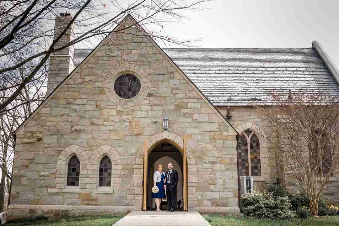 A couple stands at the entrance of a stone church building with arched windows and a peaked roof, holding hands and smiling.