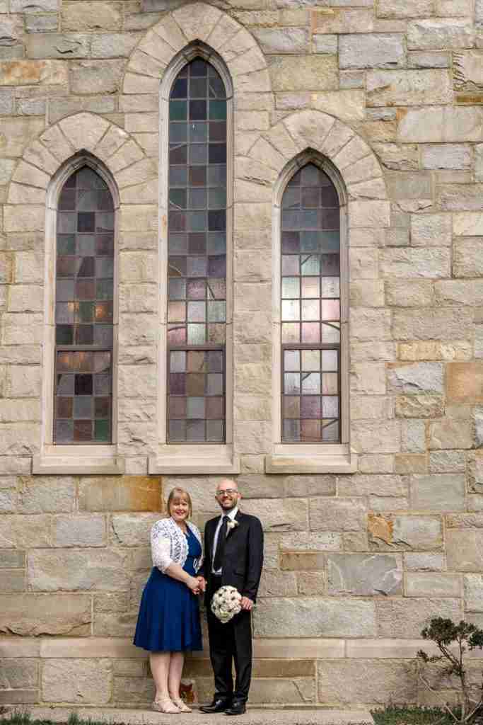 A couple dressed in semi-formal attire posing in front of a stone building with tall stained-glass windows.