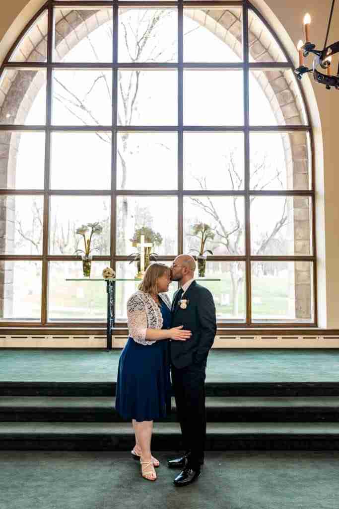 A couple shares a kiss in a church before a large arched window.