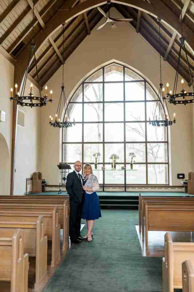 A couple standing together inside a church with large windows and wooden pews.