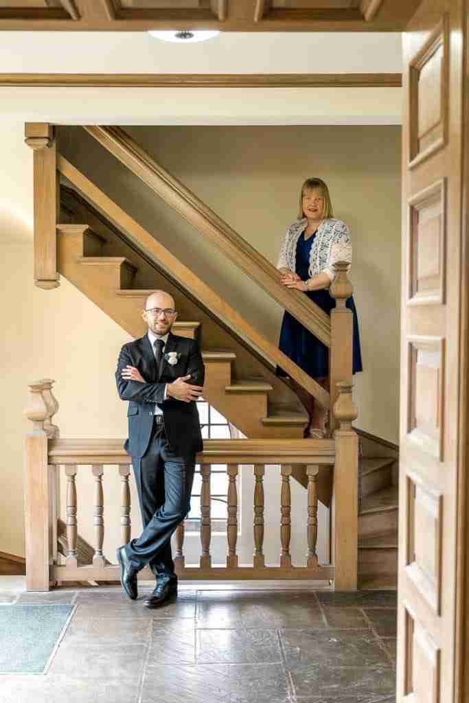 A man standing at the base of a wooden staircase with arms crossed, looking at the camera, while a woman in a blue dress stands on the landing above, also facing the camera.