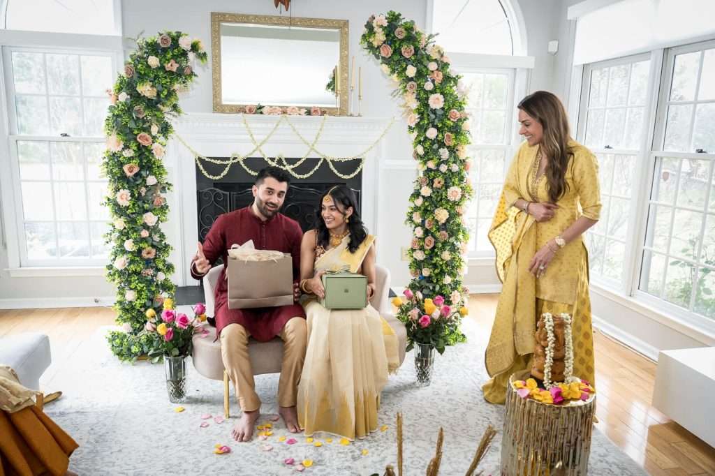 Gift Opening Scene: The couple shares a joyful moment opening gifts surrounded by lush floral arrangements, marking a memorable step towards their future together.