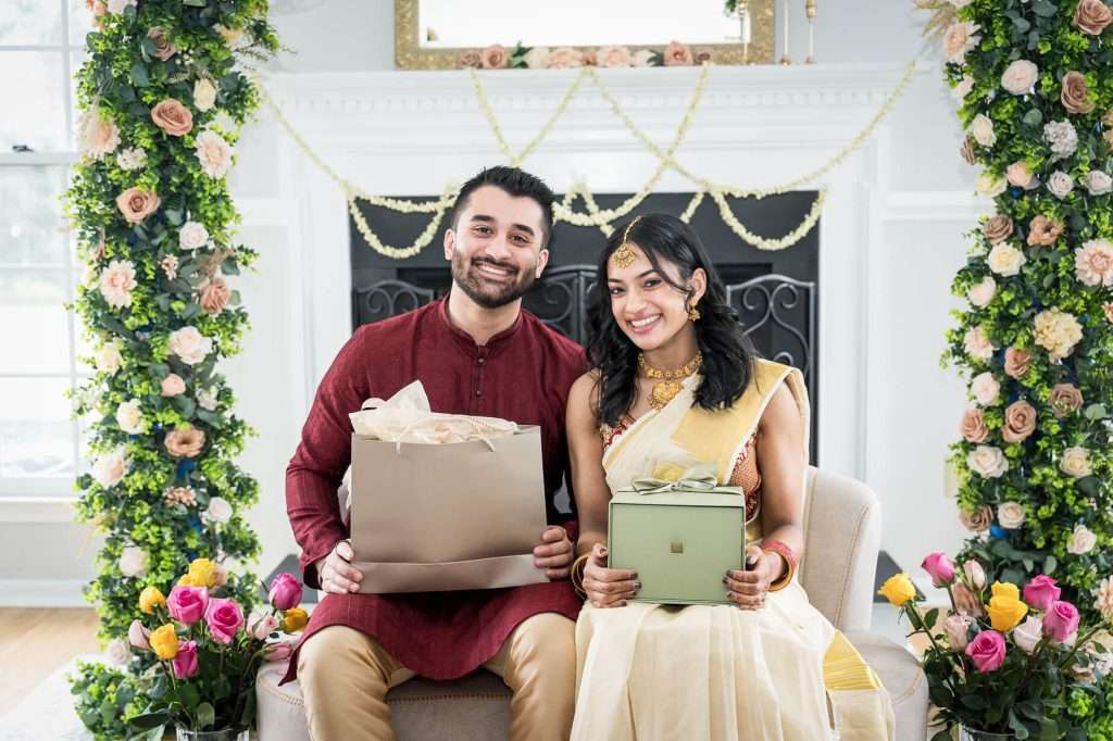 Couple With Gifts: In a setting adorned with elegant floral decor, this couple shares the excitement of opening engagement gifts, emphasizing the joy and anticipation of their upcoming wedding.