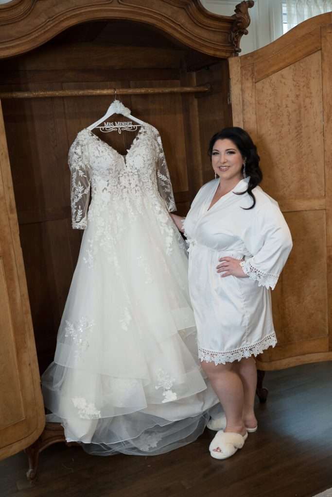 The bride, wearing a white robe, poses next to her wedding dress hanging in an antique wooden wardrobe at The Park Savoy Estate.