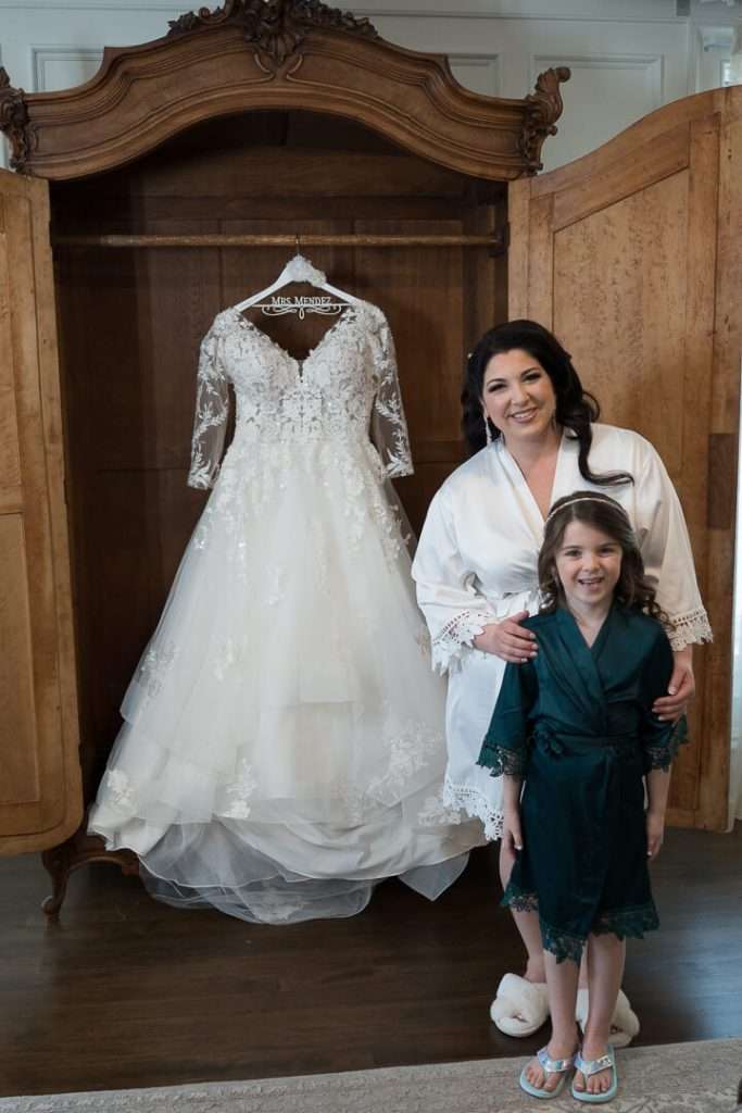 The bride, in a white robe, stands with a flower girl in a green robe in front of her wedding dress at The Park Savoy Estate.