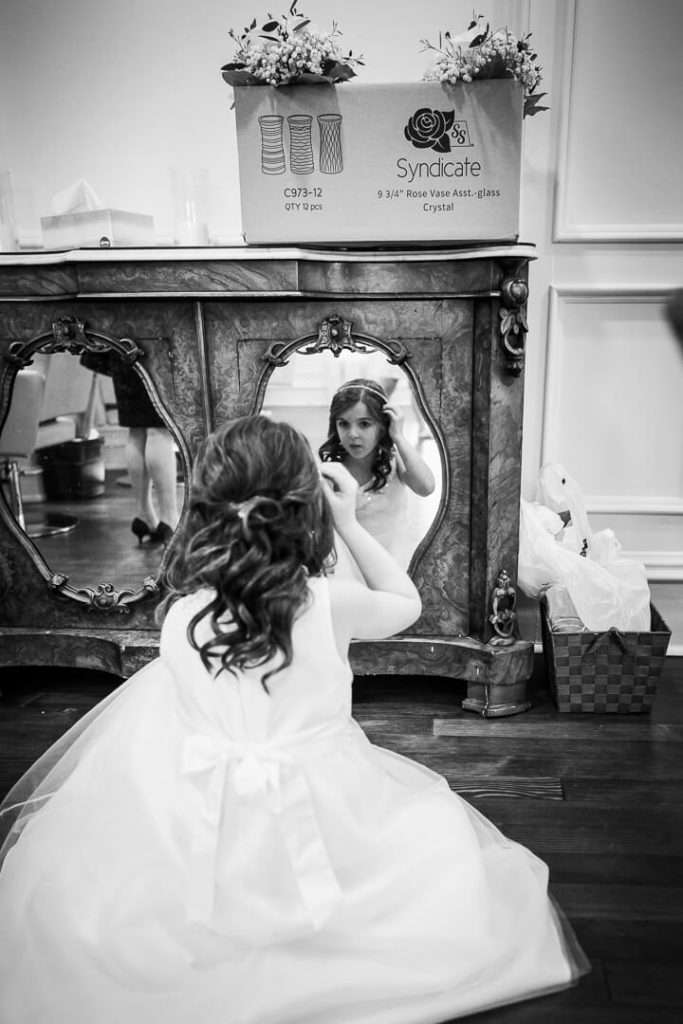 A flower girl in a white dress adjusts her hair while looking into a vintage mirror at The Park Savoy Estate.