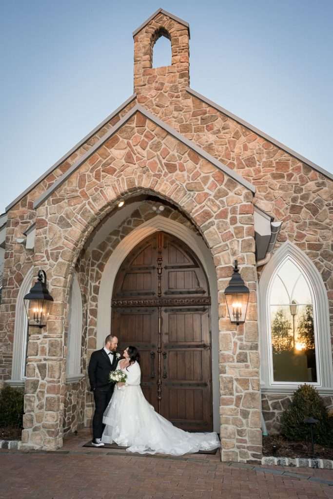 A bride and groom embrace in front of the charming stone chapel at The Park Savoy Estate, with the warm glow of sunset reflecting in the windows.
