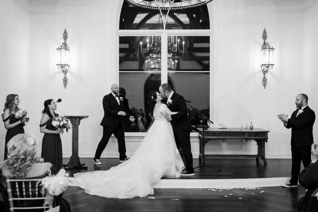 A bride and groom share their first kiss as a married couple during their wedding ceremony at The Park Savoy Estate, surrounded by their bridal party and guests.