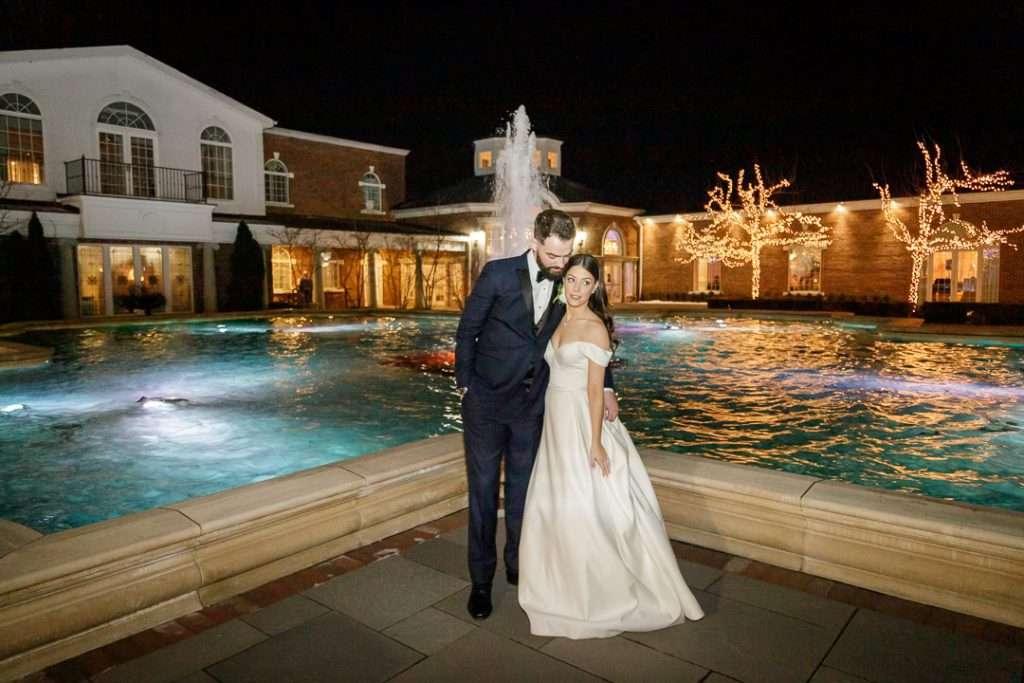 A bride and groom pose by a romantic illuminated fountain at The Rockleigh at night