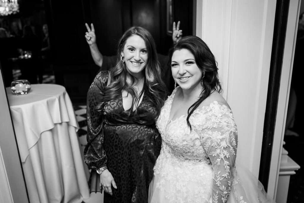 Black and white photo of the bride smiling alongside a friend at The Park Savoy, with a guest playfully making bunny ears in the background.