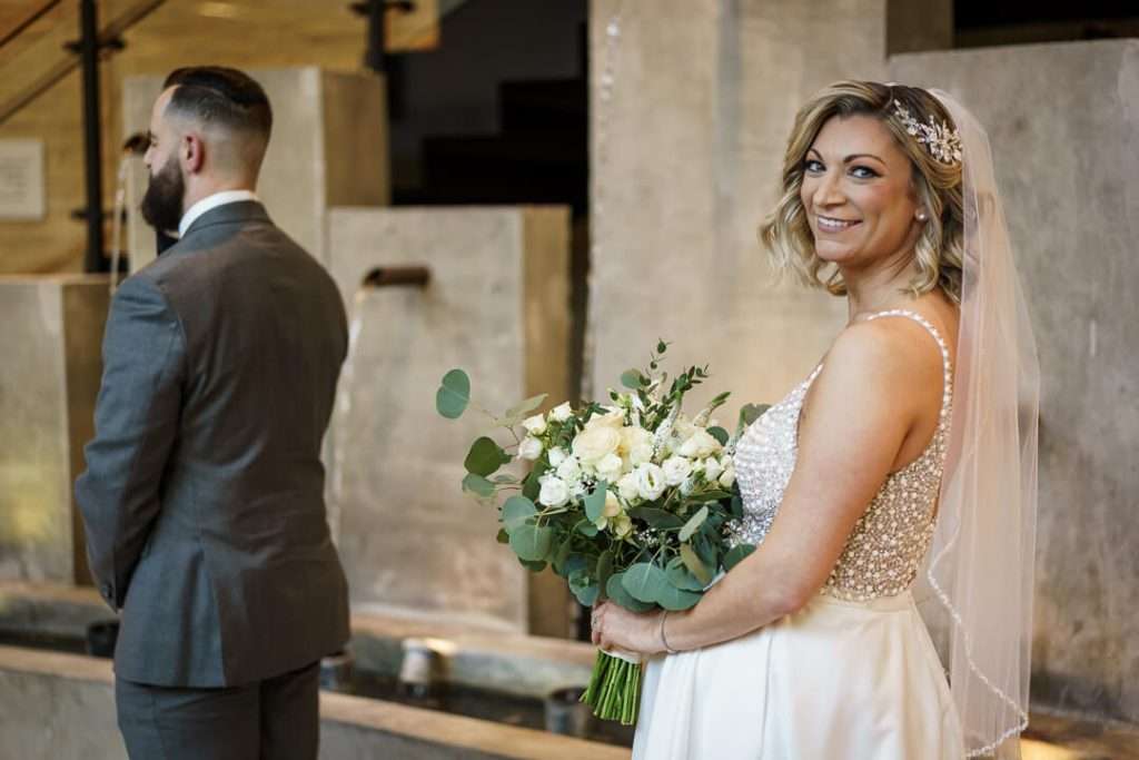 Bride smiling while holding her bouquet as the groom stands with his back turned at Stone House at Stirling Ridge.
