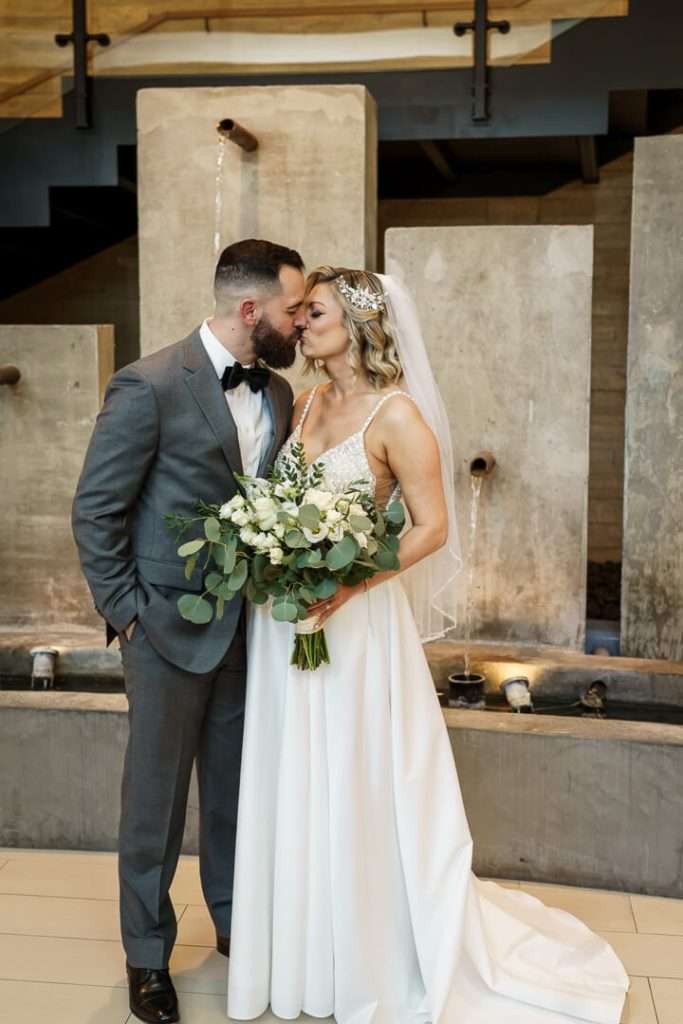 Bride and groom share their first kiss, standing in front of an elegant water feature at Stone House at Stirling Ridge.