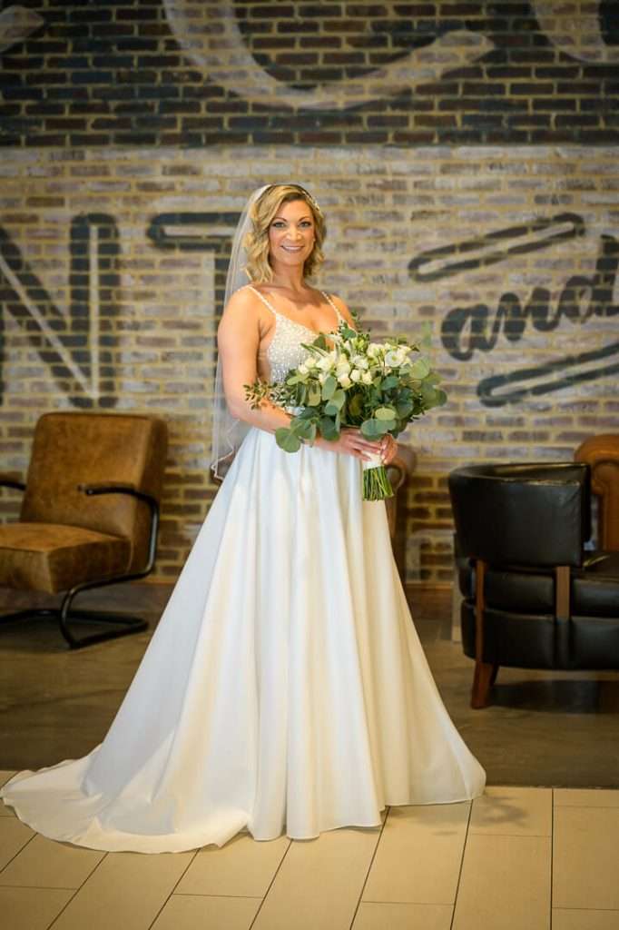 Bride posing alone, holding her bouquet, in front of a stylish brick wall at Stone House at Stirling Ridge