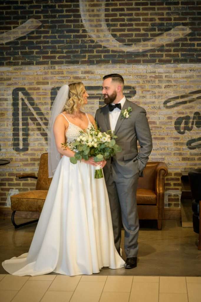 Bride and groom gazing at each other, standing in front of a stylish brick backdrop at Stone House at Stirling Ridge.