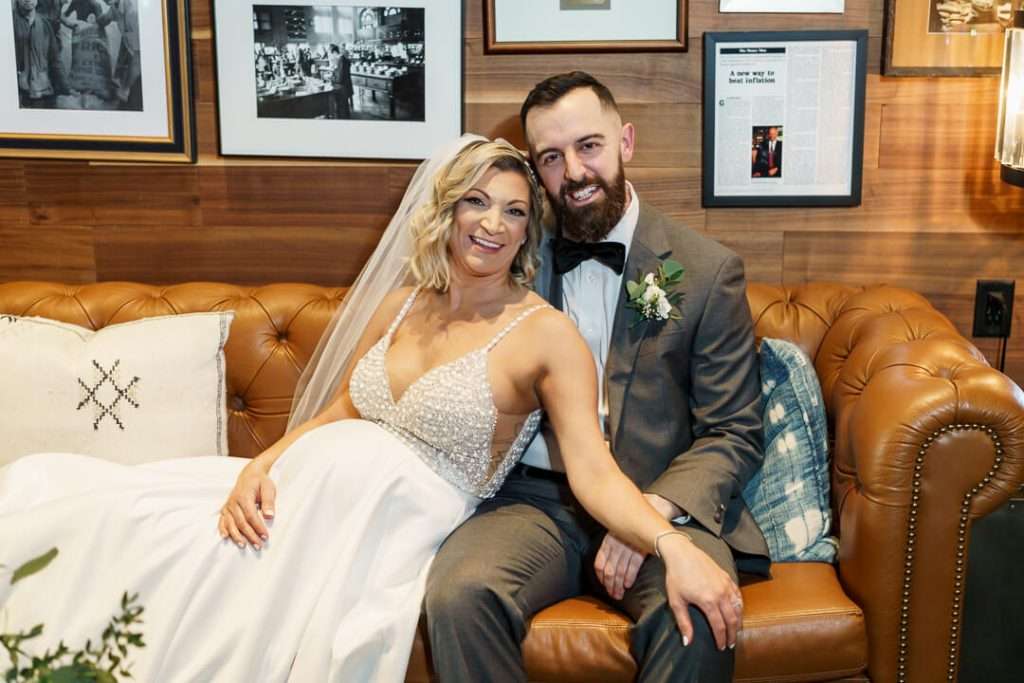 Bride and groom sitting on a stylish leather couch, smiling warmly at Stone House at Stirling Ridge