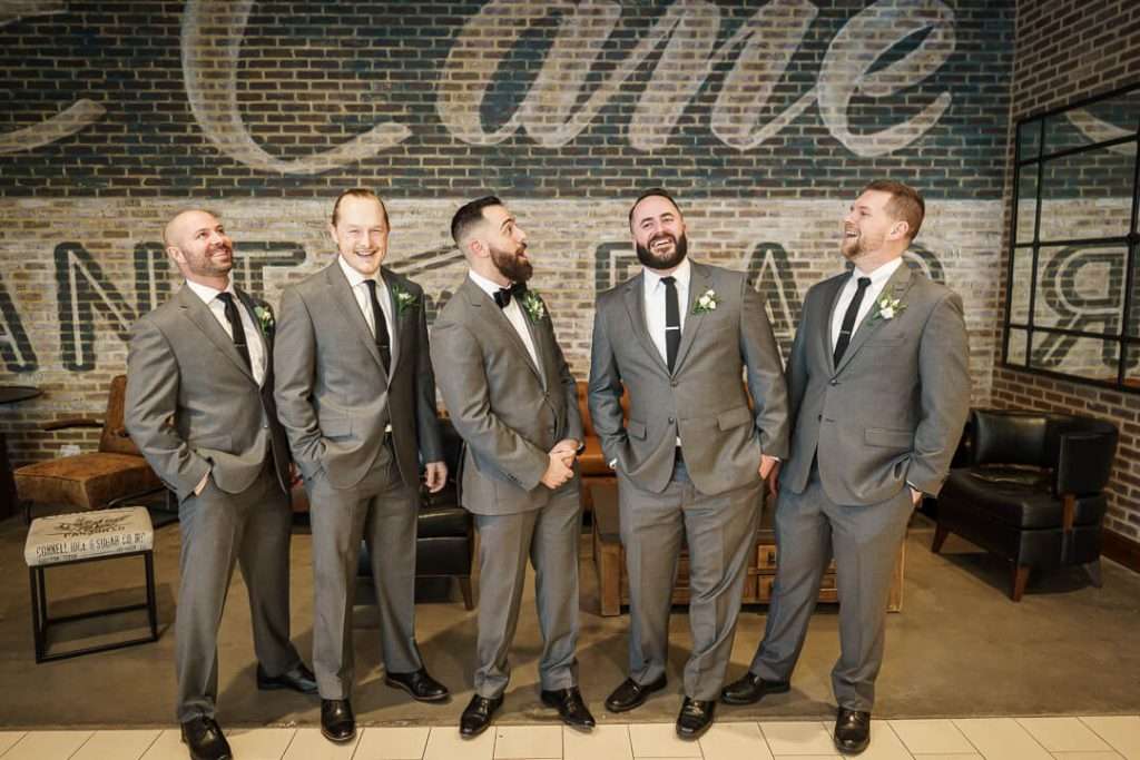 Groomsmen sharing a candid, laughing moment in front of a brick backdrop at Stone House at Stirling Ridge