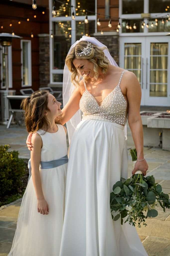 Bride sharing a sweet moment with a young flower girl outside, both smiling lovingly at each other at Stone House at Stirling Ridge