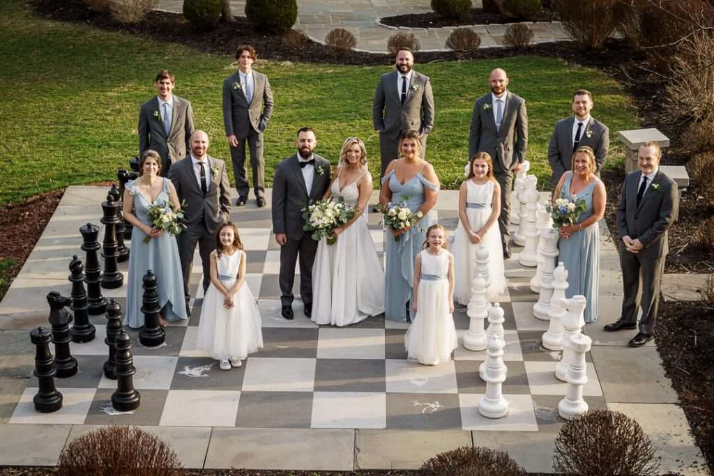 Bride and groom standing with their wedding party on a life-size chessboard outside at Stone House at Stirling Ridge