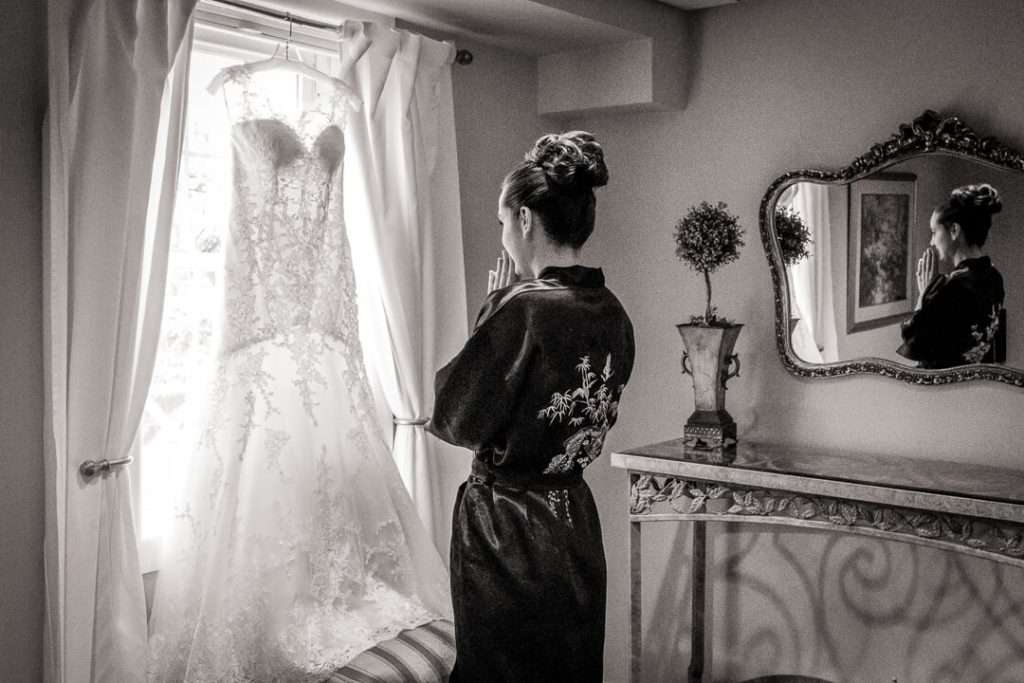 Bride in robe gazing at wedding dress hanging in window, captured in black and white by NJ wedding photographer Alex Kaplan