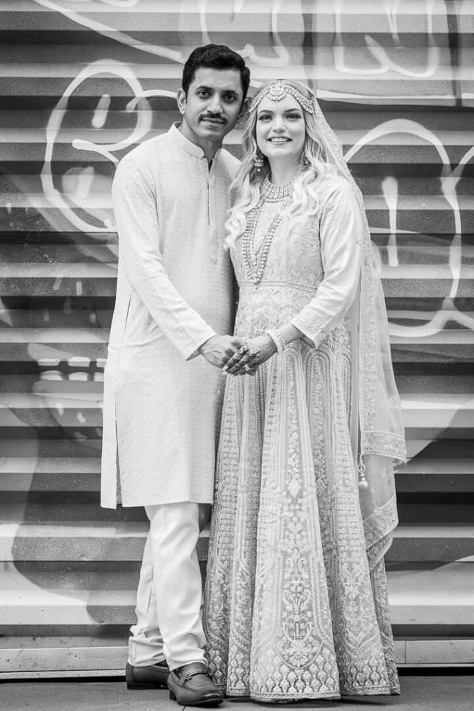 South Asian couple holding hands in traditional attire with graffiti urban wall background in black and white.
