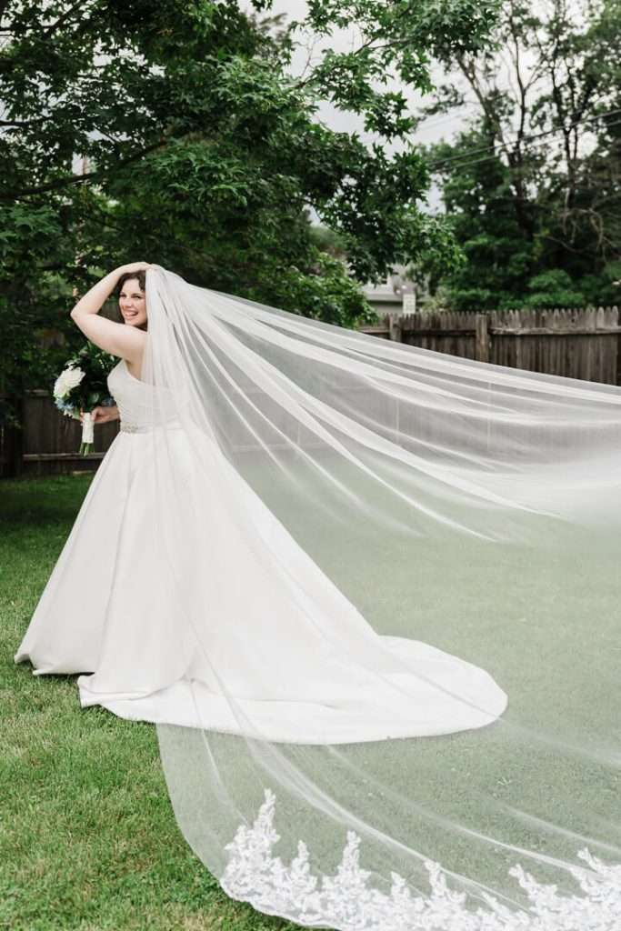 Bride smiling while holding bouquet and veil flowing dramatically behind her in a backyard in Pequannock Township, NJ.