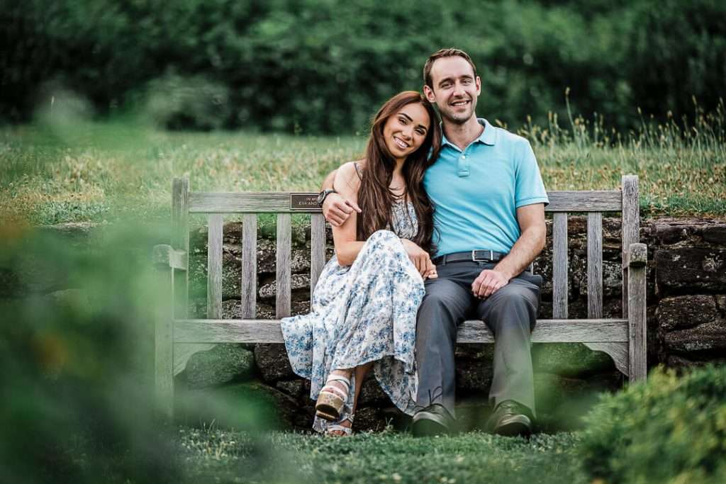 Engaged couple sitting close together on a wooden bench in Deep Cut Gardens, smiling warmly with greenery softly framing the image.