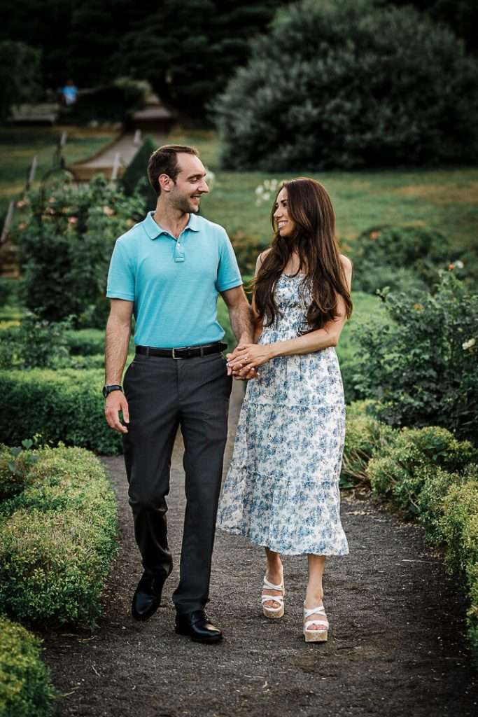 Engaged couple walking hand-in-hand through garden path at Deep Cut Gardens NJ, smiling at each other during summer engagement session