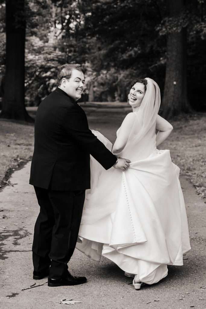 Groom lifting bride’s dress as they walk and laugh together on a tree-lined path in Nutley, NJ