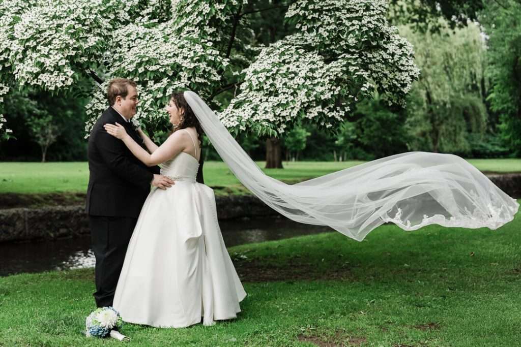 Bride and groom embracing as bride’s veil floats behind her in romantic portrait under flowering tree in Nutley, NJ