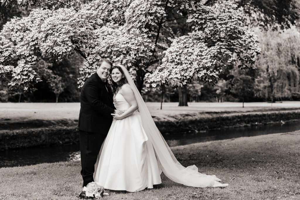 Bride and groom smiling and leaning together under blooming tree near creek in Nutley, NJ park
