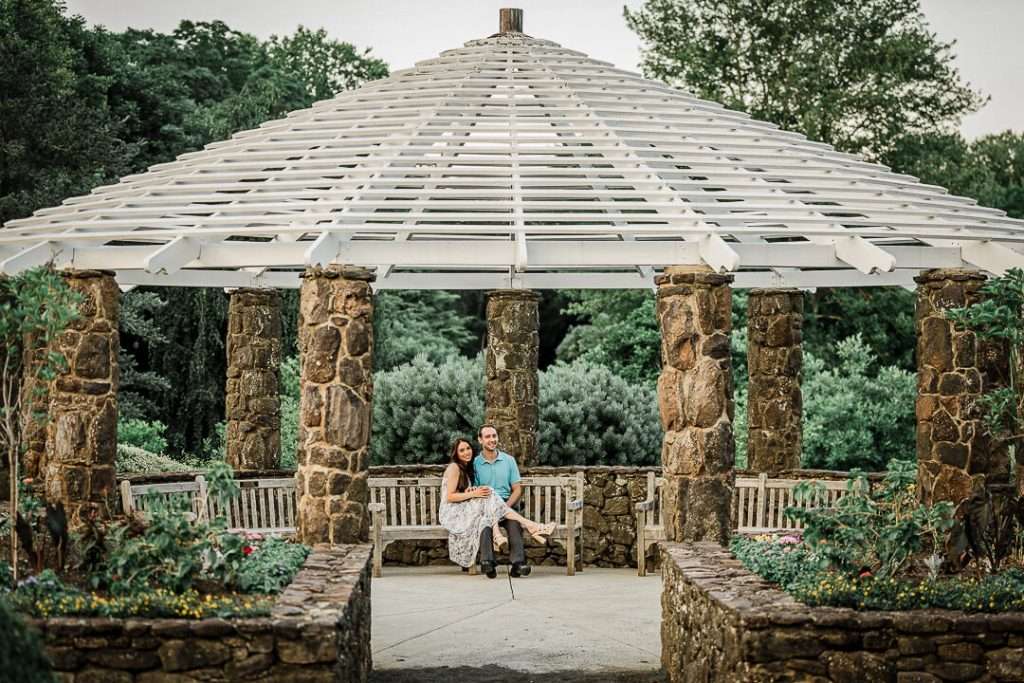 Romantic engagement portrait of a couple seated beneath the large stone-columned white pergola at Deep Cut Gardens, framed by garden flowers and greenery.