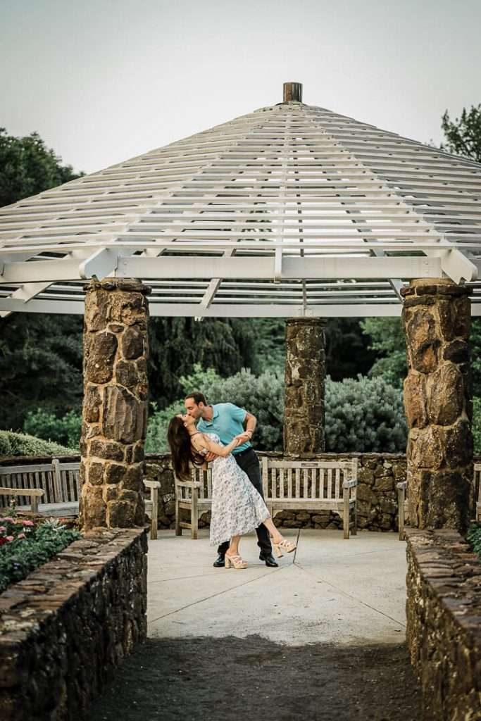 Man dipping his fiancée for a kiss beneath a white, open-beam gazebo with stone columns at Deep Cut Gardens in New Jersey.