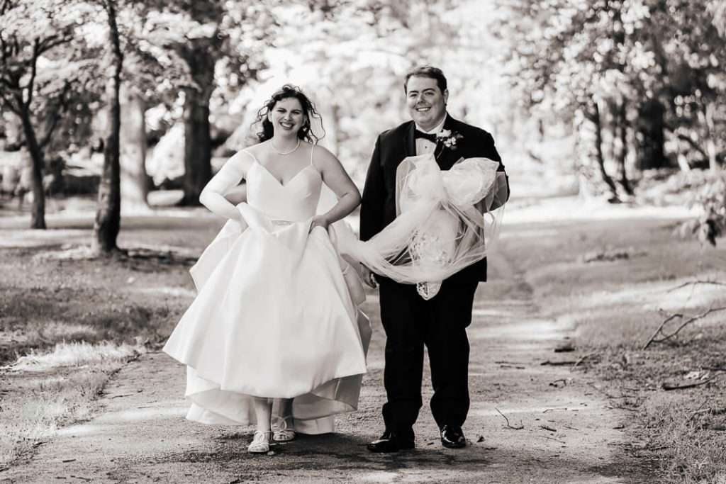 Bride and groom walking and laughing with veil in hand on a shaded trail in Nutley park