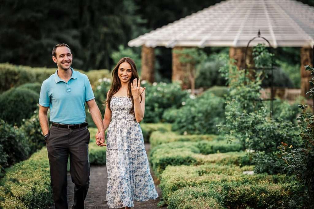 Engaged couple walking hand in hand through a lush garden path, with the woman smiling and showing off her engagement ring at Deep Cut Gardens in New Jersey.