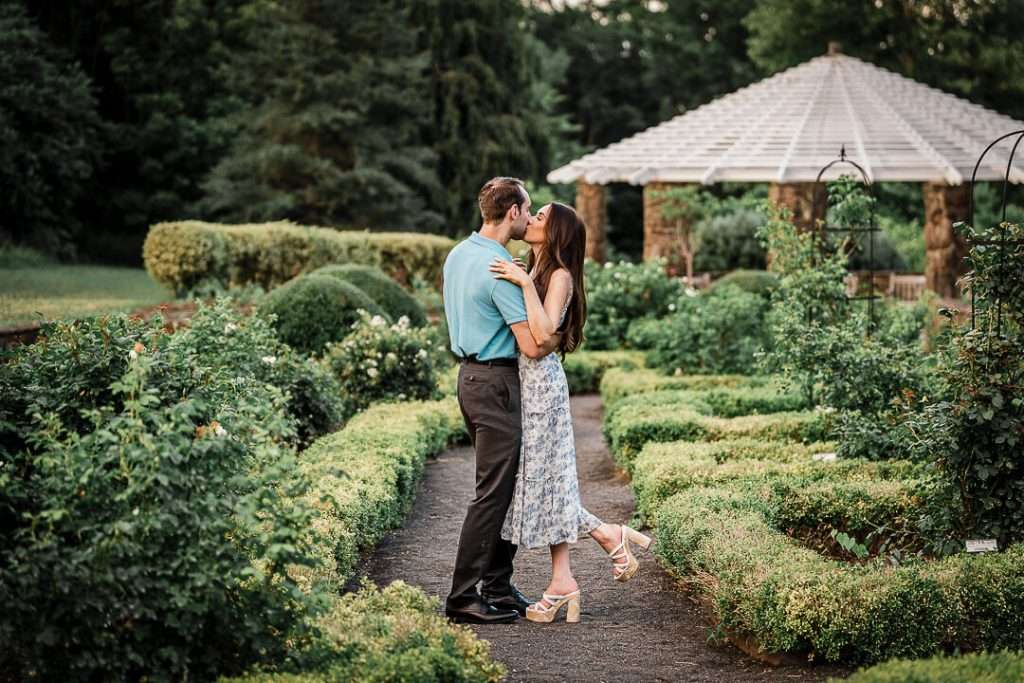 Newly engaged couple sharing a kiss on a garden path surrounded by blooming greenery and a white gazebo at Deep Cut Gardens during golden hour.