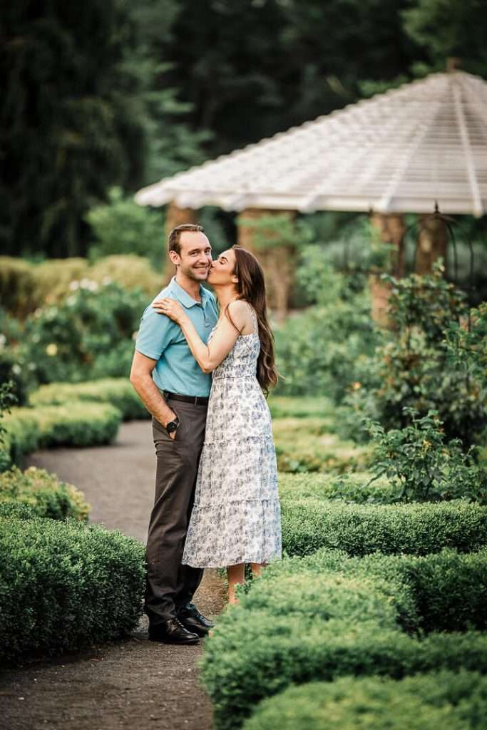 Woman kissing her smiling fiancé on the cheek while standing in a manicured garden path surrounded by lush hedges and a white gazebo in the background.