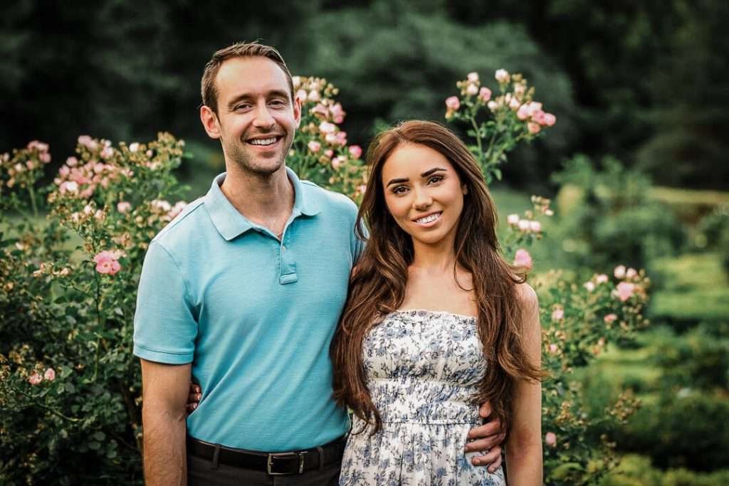 Engaged couple posing together in front of blooming rose bushes at Deep Cut Gardens, both smiling during their surprise proposal photo session in New Jersey.