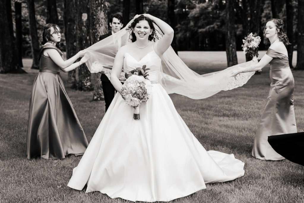 Bride laughing as bridesmaids hold her cathedral veil during portraits at Booth Park in Nutley, NJ.