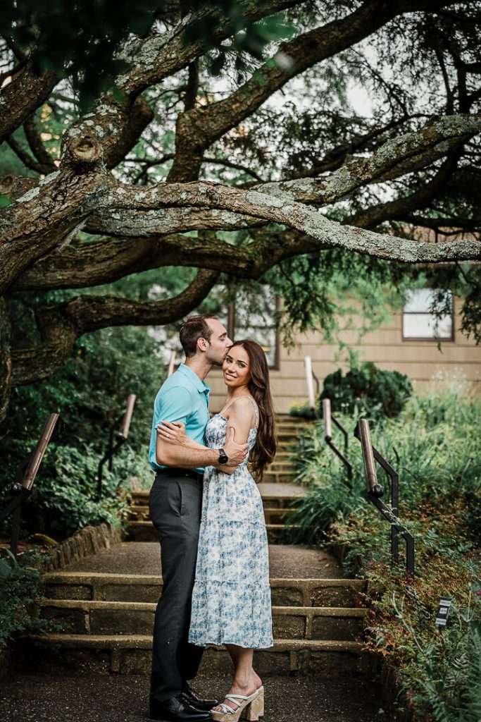Couple embracing on garden steps under a sprawling tree as the man gently kisses the woman’s forehead and she smiles at the camera.