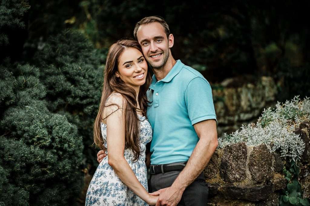 Engaged couple holding hands and smiling in a garden setting, framed by lush greenery and stone walls.