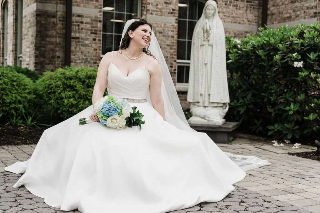 Bride seated in front of a statue at Our Lady of Good Counsel Church holding a bouquet of white and blue flowers