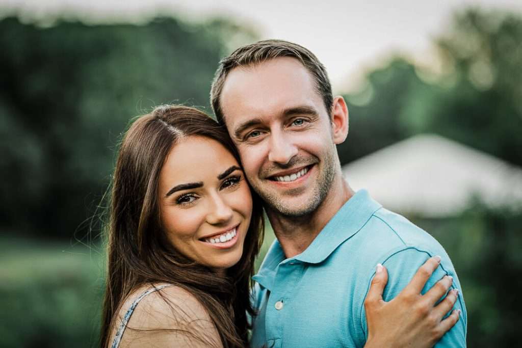 Close-up of a smiling engaged couple embracing outdoors, with greenery softly blurred in the background.
