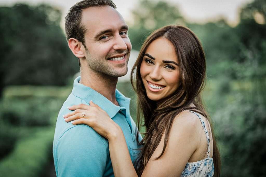 Engaged couple smiling at the camera with the woman's hand on her fiancé’s shoulder, showing off her engagement ring.