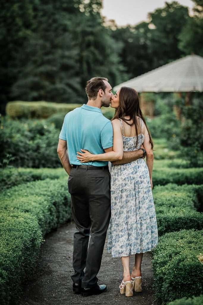 Couple sharing a kiss on a winding garden path, surrounded by lush greenery and hedges at Deep Cut Gardens.
