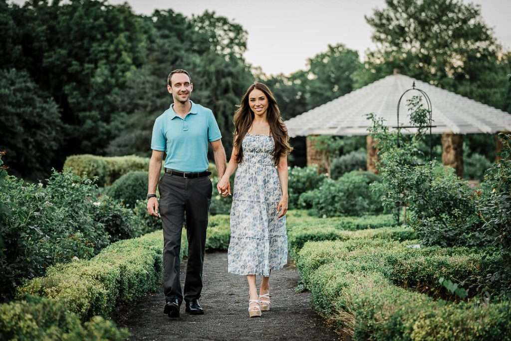 Couple walking hand-in-hand through a beautifully manicured garden path at Deep Cut Gardens, surrounded by lush greenery and trimmed hedges.

