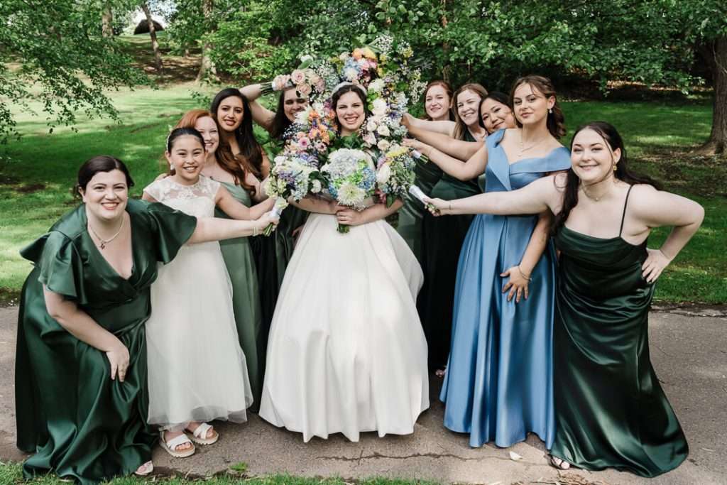 Bride surrounded by smiling bridesmaids holding bouquets in a flower frame formation at Booth Park