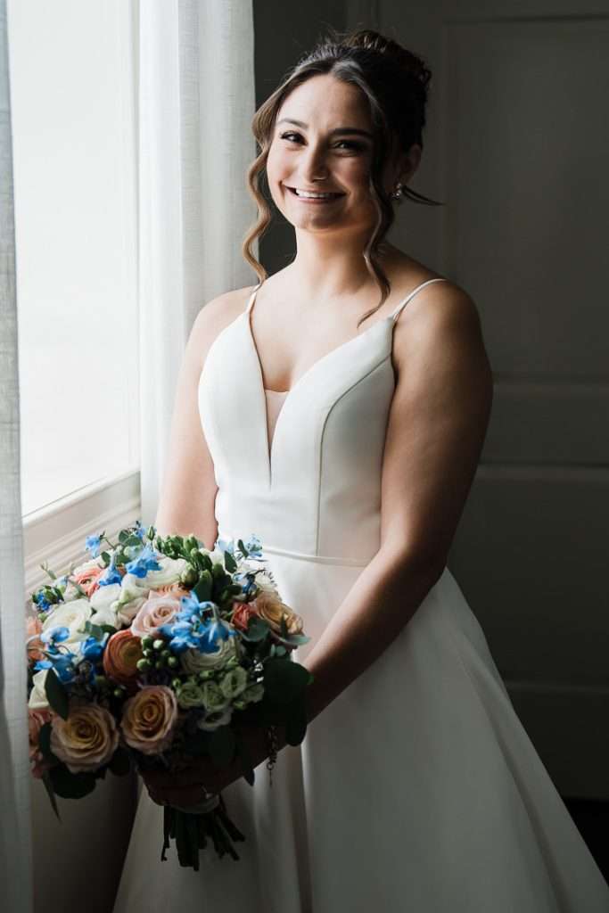 Bride stands by a window in natural light, holding a bouquet of pastel and blue flowers, smiling in her wedding dress at The Mansion at Mountain Lakes.