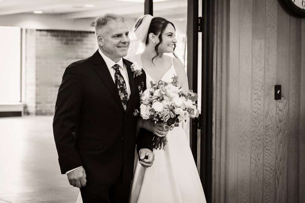 Bride enters the church on her father’s arm, both smiling as they walk down the aisle with a colorful bouquet at Notre Dame of Mt. Carmel Roman Catholic Church.