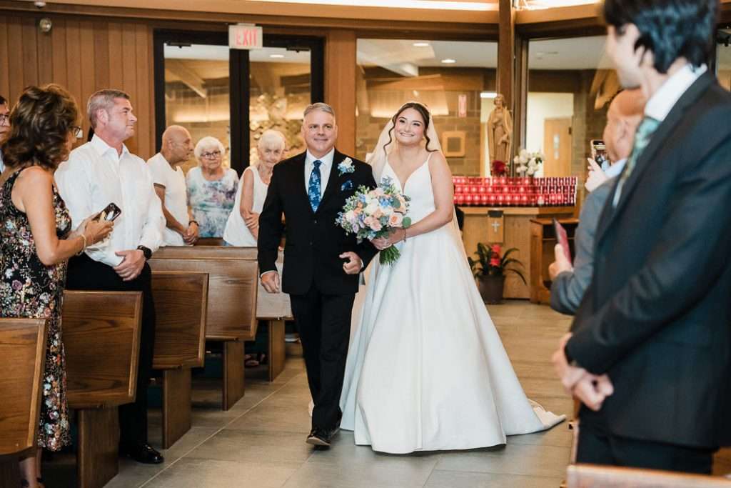 Bride walks down the aisle arm-in-arm with her father, smiling warmly and holding a pastel bouquet at Notre Dame of Mt. Carmel Roman Catholic Church.